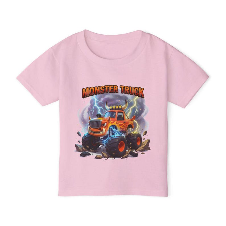 Puede incluir: Una camiseta rosa claro con un dise&ntilde;o de cami&oacute;n monstruo de dibujos animados. El cami&oacute;n naranja tiene neum&aacute;ticos negros grandes con llantas rojas, sobre un fondo de rel&aacute;mpagos y rocas. Las palabras "MONSTER TRUCK" est&aacute;n impresas encima del cami&oacute;n.