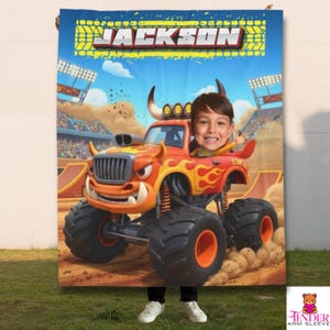 Op de afbeelding: Een gepersonaliseerde deken met een cartoon monstertruck met het gezicht van een kind achter het stuur. De truck is oranje en rood met vlammen, tegen een blauwe lucht en een zandbaan. De naam "JACKSON" staat bovenaan.