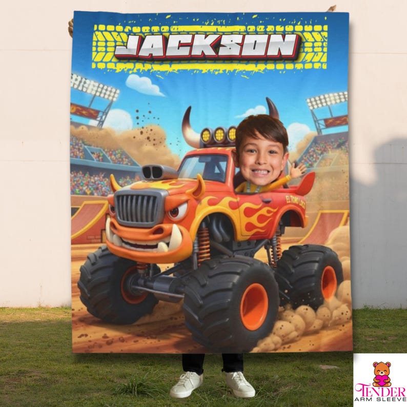 Op de afbeelding: Een gepersonaliseerde deken met een cartoon monstertruck met het gezicht van een kind achter het stuur. De truck is oranje en rood met vlammen, tegen een blauwe lucht en een zandbaan. De naam "JACKSON" staat bovenaan.