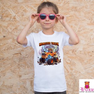 Puede incluir: Una camiseta blanca con un dise&ntilde;o de cami&oacute;n monstruo colorido y el texto "MONSTER TRUCK". El gr&aacute;fico incluye un cami&oacute;n naranja con llamas, rayos y neum&aacute;ticos grandes. El ni&ntilde;o lleva gafas de sol rosas.