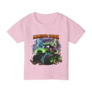 May include: A light pink t-shirt featuring a cartoon monster truck design. The monster truck is green and purple with glowing green wheels, set against a backdrop of lightning and clouds. The words "MONSTER TRUCK" are above the truck.