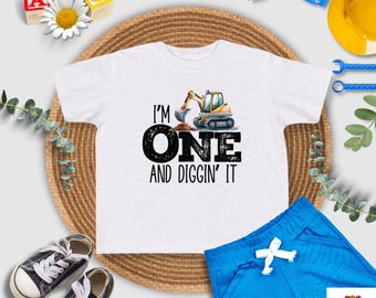 May include: White short-sleeved t-shirt with a yellow excavator graphic and the text "I'm One and Diggin' It".