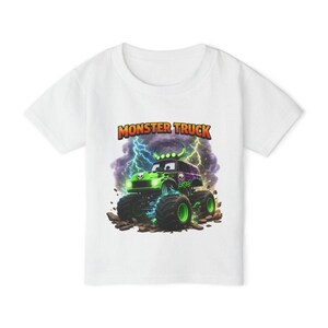 May include: White t-shirt featuring a colorful graphic of a monster truck with a skull design, lightning bolts, and the words "Monster Truck" in orange and yellow. The truck is green and purple.