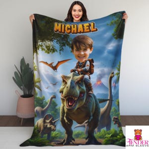 May include: A personalized blanket featuring a cartoon illustration of a boy riding a Tyrannosaurus Rex in a prehistoric landscape. The blanket has the name "MICHAEL" in large orange letters at the top.