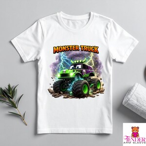 May include: White t-shirt featuring a colorful monster truck graphic. The monster truck is green and purple with large black tires and a skull design. The text "MONSTER TRUCK" is displayed above the truck in orange and yellow.