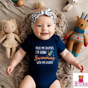 Puede incluir: Un bebé con un body azul marino con el texto "Pack my diapers I'm going swimming with my daddy!" y un gráfico de piña. El bebé lleva una diadema blanca y negra y sonríe.