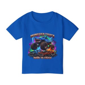 May include: A royal blue t-shirt featuring a colorful graphic of a monster truck with a shark-like design, jumping over crushed cars. The text "MONSTER TRUCK" and "BORN TO CRUSH" are displayed above and below the truck.