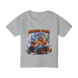 Puede incluir: Una camiseta gris con un dise&ntilde;o de cami&oacute;n monstruo de dibujos animados. El cami&oacute;n naranja tiene neum&aacute;ticos negros grandes y est&aacute; sobre un fondo de rel&aacute;mpagos y rocas. Las palabras "MONSTER TRUCK" est&aacute;n impresas en naranja encima del cami&oacute;n.