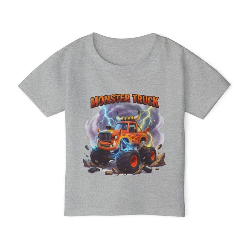 Puede incluir: Una camiseta gris con un dise&ntilde;o de cami&oacute;n monstruo de dibujos animados. El cami&oacute;n naranja tiene neum&aacute;ticos negros grandes y est&aacute; sobre un fondo de rel&aacute;mpagos y rocas. Las palabras "MONSTER TRUCK" est&aacute;n impresas en naranja encima del cami&oacute;n.