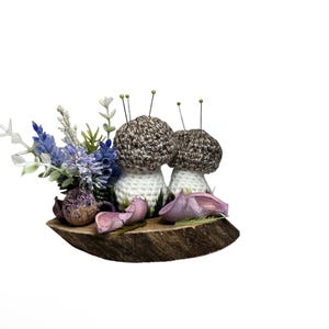 May include: Two crocheted mushrooms with brown and white yarn, sitting on a wooden slice with dried lavender and pink flower petals.
