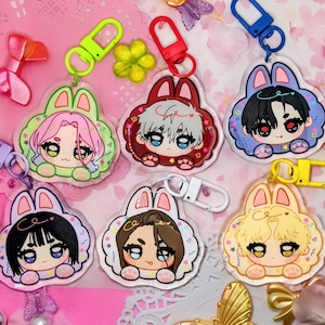 May include: A collection of anime-style keychains featuring characters with bunny ears. Each keychain has a different hair color and outfit, with a clear acrylic design and colorful accents. The keychains are attached to various colored clasps.