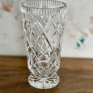 May include: A clear, cut-glass vase with a diamond pattern and a round base. The vase has a cylindrical shape with vertical lines at the rim. The glass reflects light, creating a sparkling effect. The vase is likely used for displaying flowers or as a decorative item.
