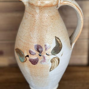 May include: A white ceramic pitcher with a brown glaze and a floral design in shades of brown, pink, and green. The pitcher has a handle and a spout.