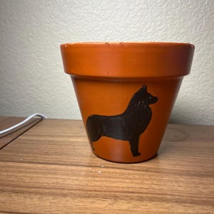 Pode incluir: Um vaso de terracota com uma silhueta preta de um cão pintada no lado.