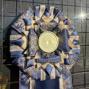 May include: A circular award rosette featuring layered navy blue and gold glitter ribbon with snowflake patterns. A central gold medallion displays the text American Kennel Club Founded 1884. Three long ribbon tails extend from the base.
