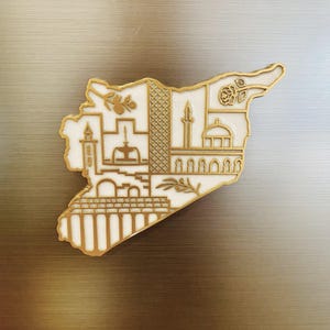 May include: A gold and white metal pin in the shape of Syria. The pin features detailed illustrations of landmarks and symbols, including a rose, mosque, and olive branch. The pin is set against a metallic background.