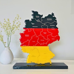 May include: A decorative map of Germany in the shape of the country, featuring the German flag colors: black, red, and yellow. The map is divided into regions with their names labeled. It is mounted on a black base and placed on a white surface.