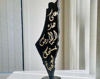 May include: A vertical black sculpture shaped like a map of Palestine, featuring gold Arabic calligraphy script running down the center. The piece is mounted on a circular black base, creating a smooth, glossy finish with a distinct silhouette.