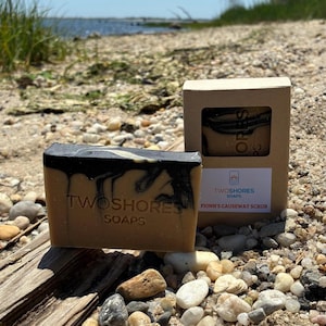 May include: A bar of soap with a dark top and light brown base, with the text "TWOSHORES SOAPS" embossed on the front. A box of soap with the text "FIONN'S CAUSEWAY SCRUB" is also visible. The scene is set on a beach.