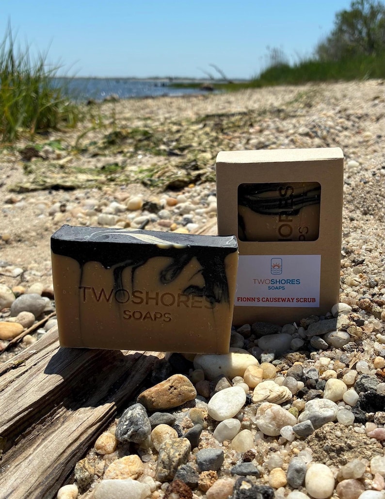 May include: A bar of soap with a dark top and light brown base, with the text "TWOSHORES SOAPS" embossed on the front. A box of soap with the text "FIONN'S CAUSEWAY SCRUB" is also visible. The scene is set on a beach.