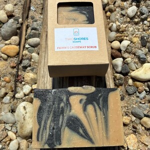 May include: A bar of soap with a swirling black and tan design, next to its open cardboard box. The box has a label that reads "TWOSHORES SOAPS" and "FIONN'S CAUSEWAY SCRUB". The soap is on a piece of driftwood, surrounded by pebbles.