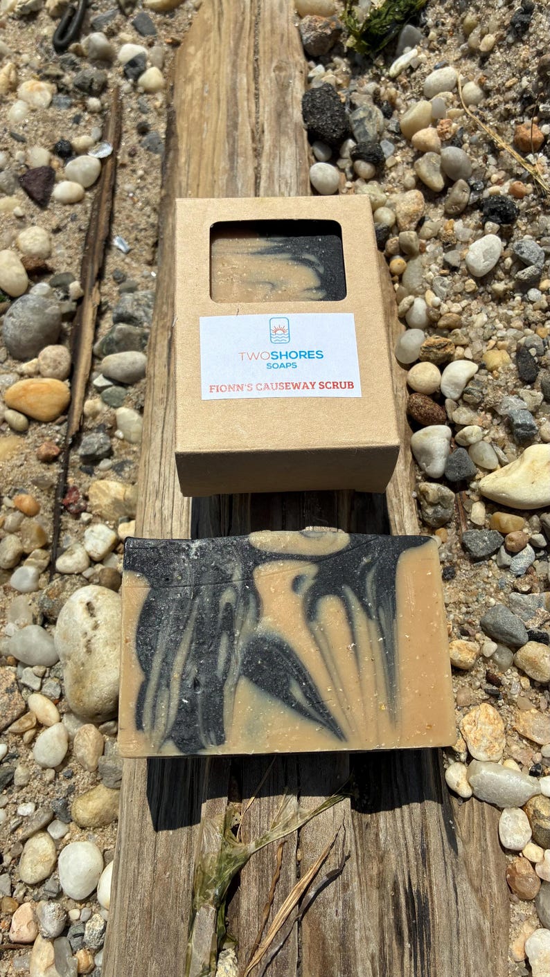 May include: A bar of soap with a swirling black and tan design, next to its open cardboard box. The box has a label that reads "TWOSHORES SOAPS" and "FIONN'S CAUSEWAY SCRUB". The soap is on a piece of driftwood, surrounded by pebbles.