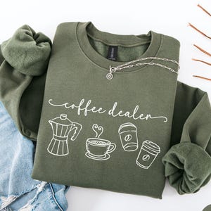 May include: Olive green sweatshirt with the text "coffee dealer" and coffee-related graphics. The design includes a coffee pot, a steaming cup, and two to-go coffee cups. A silver necklace is draped on the sweatshirt. The sweatshirt is paired with blue jeans.
