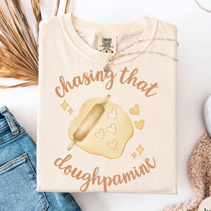 May include: Ivory-colored t-shirt with the text "chasing that doughpamine" in a copper color. The graphic features a rolling pin and dough with heart cutouts. The shirt is folded with a pair of blue jeans and a necklace.