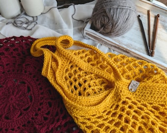 Può includere: Una borsa tote all'uncinetto gialla fatta a mano, a trama aperta, con una lunga tracolla, accanto a un centrino all'uncinetto bordeaux. Un gomitolo di filato grigio, uncinetti e altri materiali artigianali sono sullo sfondo. La borsa ha una piccola etichetta.
