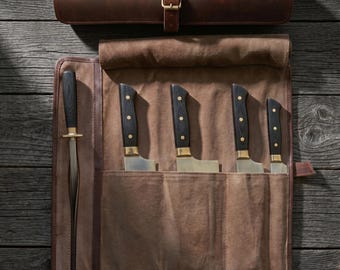 May include: A brown leather knife roll with a matching leather roll-up case. The roll contains five knives with black handles, brass bolsters, and silver blades. A honing steel is also included. The set is displayed on a wooden surface.