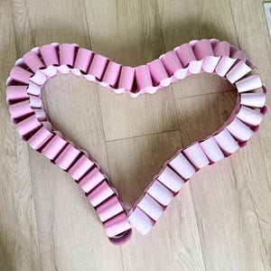 May include: A heart-shaped decoration constructed from a continuous chain of interlocking light pink paper loops. The paper links are arranged in a uniform circular pattern to form a large, open heart shape with a smooth, consistent texture.