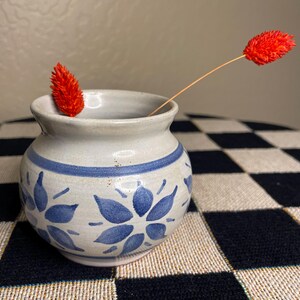 Puede incluir: Un pequeño jarrón de cerámica blanca con un patrón floral azul. El jarrón está sobre una superficie a cuadros en blanco y negro. Una sola flor roja está en el jarrón.