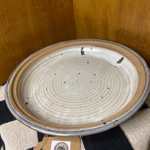 Puede incluir: Un plato de cerámica hecho a mano con un centro color crema y un borde marrón. El plato tiene un diseño en espiral y un borde exterior gris. Una etiqueta marrón con un logotipo está adjunta al plato.