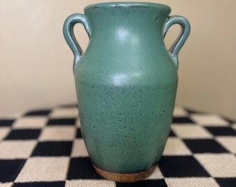 May include: A small, green ceramic vase with two curved handles. The vase has a speckled texture and sits on a small, round, cork base. The background is a neutral color.