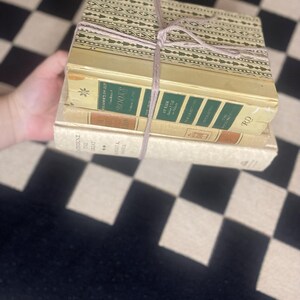 Puede incluir: Una pila de libros antiguos atados con hilo. Los libros tienen cubiertas y lomos de color crema con letras verdes y doradas. El libro superior tiene una cubierta estampada decorativa en tonos verdes y dorados.