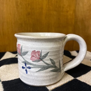 May include: A white ceramic mug with a floral design. The mug has a speckled glaze and features pink flowers, green leaves, and blue accents.