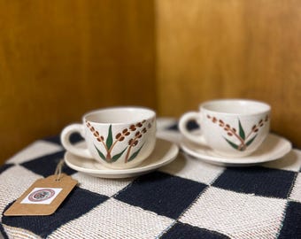 Puede incluir: Dos tazas de té y platillos de cerámica blanca con un diseño floral marrón y verde. Las tazas tienen asas y están colocadas sobre una manta a cuadros blancos y negros. Una etiqueta de precio está adjunta a la manta.