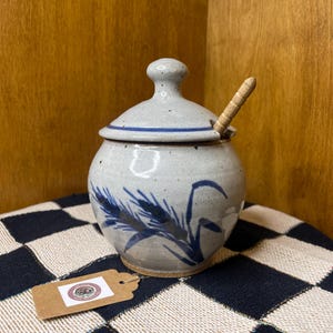 May include: A ceramic sugar bowl with a lid and a small wooden spoon. The bowl is light gray with blue wheat stalk designs. The lid has a blue stripe around the edge and a small knob. The bowl sits on a black and white checkered cloth.