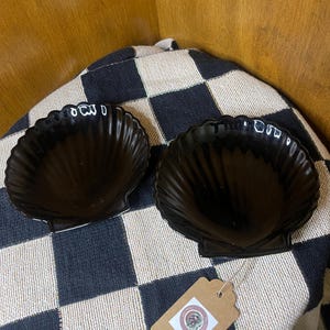 Puede incluir: Dos platos negros en forma de concha con acabado brillante. Los platos están colocados sobre un paño a cuadros blanco y negro. Una pequeña etiqueta marrón con un logotipo está adherida al paño.