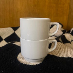 Puede incluir: Dos tazas de café de cerámica blanca apiladas una encima de la otra. Cada taza tiene un asa y un diseño cilíndrico simple. Las tazas están sobre un patrón de cuadros blancos y negros.