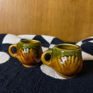 May include: Two small brown and green ceramic mugs with a textured design. The mugs have a rounded shape and a small handle.