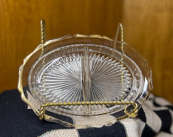 May include: A clear, oval-shaped glass serving dish with a divided interior, set in a gold-toned metal stand. The dish features a starburst pattern in the center. The stand has a twisted rope design and a scalloped edge.