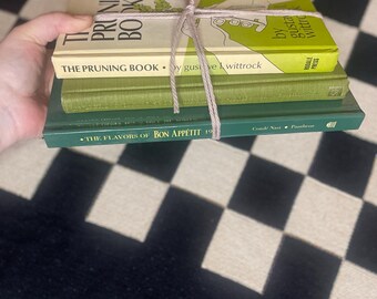 Könnte beinhalten: Ein Stapel von vier Büchern, die mit Bindfaden zusammengebunden sind. Das oberste Buch trägt den Titel "THE PRUNING BOOK" mit einem grün-weißen Einband. Das zweite Buch trägt ebenfalls den Titel "THE PRUNING BOOK". Das dritte Buch ist grün, und das untere Buch trägt den Titel "THE FLAVORS OF BON APPÉTIT 19".