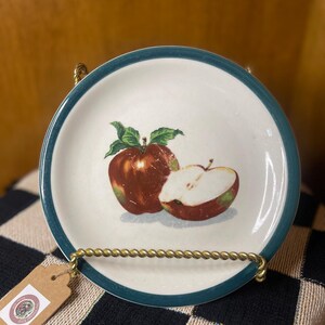 Puede incluir: Plato pequeño y redondo de cerámica con centro blanco y borde azul turquesa. Presenta una ilustración detallada de dos manzanas, una entera y otra cortada, con hojas verdes. El plato se exhibe sobre un soporte de alambre dorado.