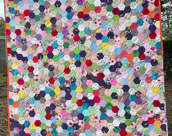 May include: A handmade quilt with a vibrant patchwork design. The quilt features numerous hexagonal fabric pieces in a variety of colors and patterns, including floral, polka dots, and solid hues. The quilt is bordered with orange fabric and is hanging outdoors.