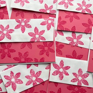 May include: A collection of pink and white stationery. The white envelopes feature a repeating pattern of pink floral designs. The background is a solid pink colour, with the same floral pattern. The stationery is arranged in a layered, overlapping fashion.