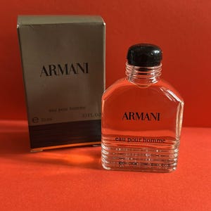 Könnte beinhalten: Eine Miniaturflasche Armani Eau pour Homme Eau de Cologne mit schwarzem Verschluss. Die transparente Glasflasche enthält eine rötliche Flüssigkeit und steht neben einer passenden Schachtel. Auf Schachtel und Flasche ist das Armani-Logo abgebildet.
