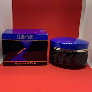 Può includere: Una scatola nera e blu con il testo "CLANDESTINE" e "Guy Laroche Paris" su di essa.  Accanto alla scatola c'è un barattolo nero e blu con un coperchio dorato. Il barattolo ha un motivo geometrico.