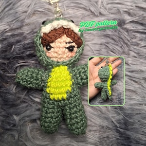 May include: Crocheted dinosaur keychain with a green body and yellow belly. The keychain features a small doll wearing a dinosaur costume with brown hair and a light pink face. The text "PDF pattern by Jaenerys crafts" is visible in the image.