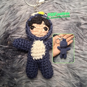 May include: A handmade crochet doll keychain in a blue dinosaur costume. The doll has a light skin tone face with black hair and large eyes. The keychain includes a metal ring. The text "PDF pattern by Jaenel's Crafts" is visible.
